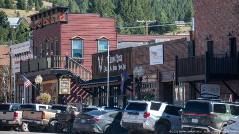 Historic Philipsburg storefronts with an upper wooden balcony, decorative window trim, business signs, awnings, and parked cars.