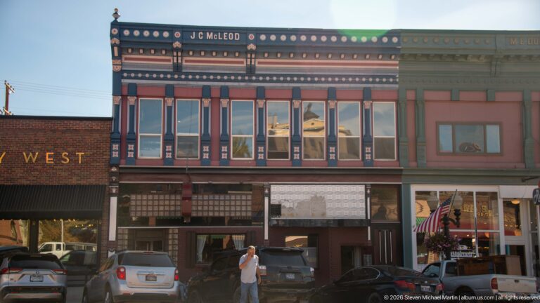 Front view of the historic J.C. McLeod Building with ornate painted cornice, tall upper windows, storefronts, vehicles, and pedestrian.