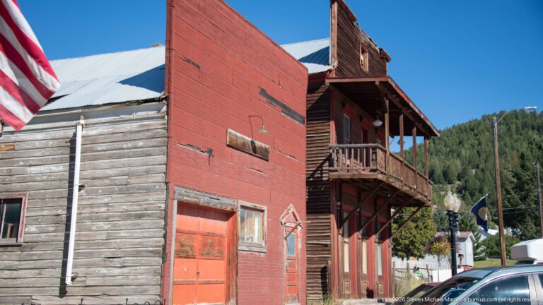 Weathered wood and red-painted rear commercial buildings with garage doors, exterior balcony, parked vehicle, and wooded mountain behind.