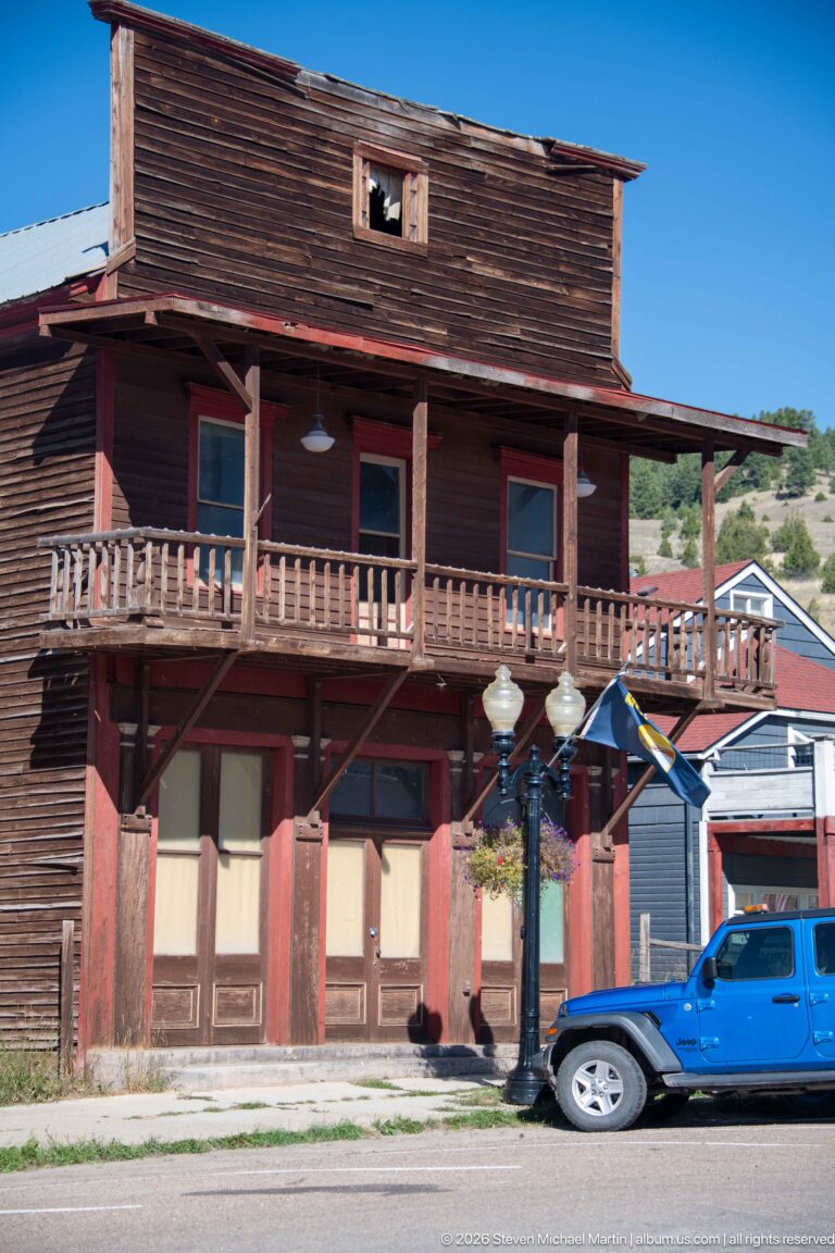 Weathered two-story wood commercial building with full-width balcony, faded red posts, boarded openings, and blue pickup at curb.