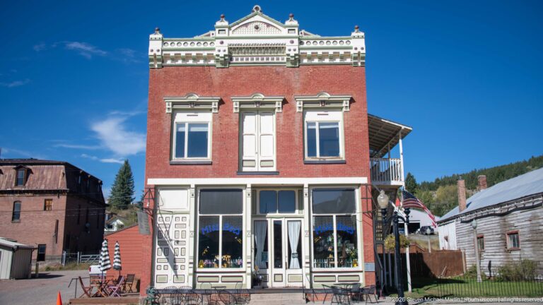 Narrow red-brick commercial building with ornate white cornice, shaped parapet, three upper bays, decorated storefront, and outdoor tables.
