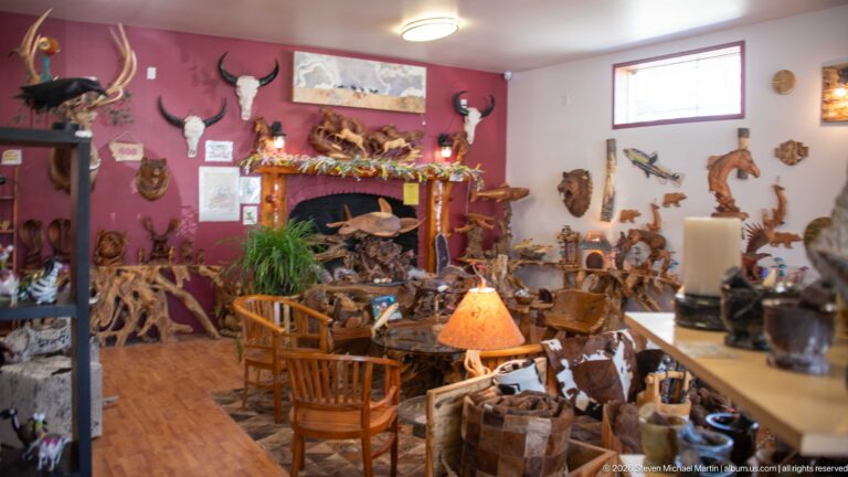 Rustic shop interior crowded with carved wooden animals, antlers, western décor, furniture, lamps, and a fireplace mantel.