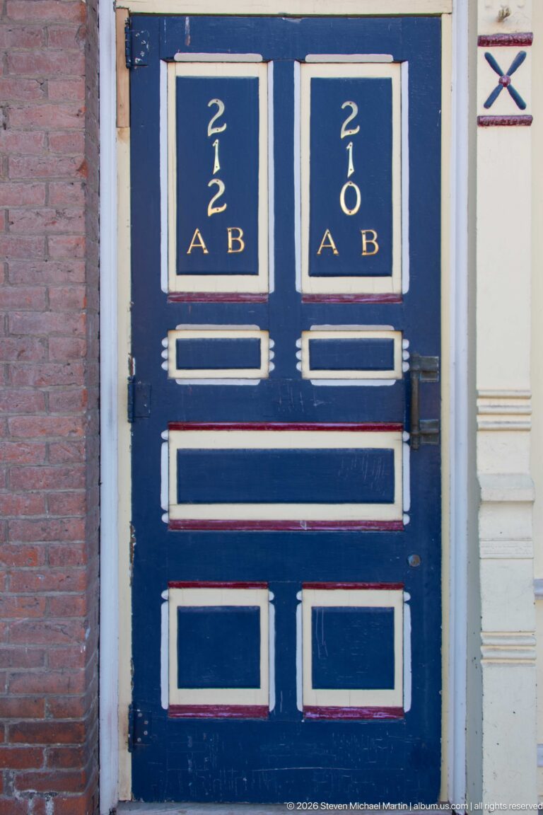 Blue double doors with cream trim, gold 212 A and 212 B numbering, rectangular panels, and brick wall at left.