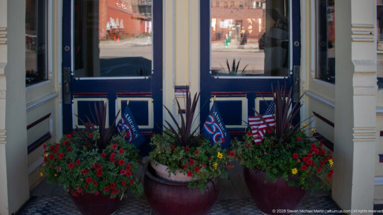 Recessed blue-and-cream storefront entrance framed by large red-flower planters, American flags, patriotic pennants, and street reflections.
