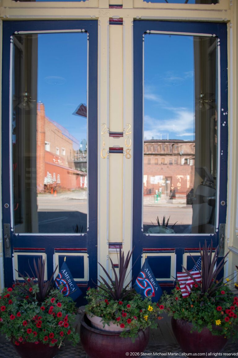 Blue-framed storefront windows above red-flower planters with American flags, reflecting brick commercial buildings across the street.