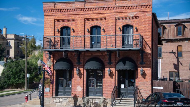 Two-story red-brick building with three arched storefront bays, tall upper openings, iron balcony, and stone base.