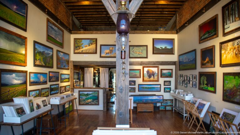 Long gallery interior filled with framed western landscapes and wildlife paintings beneath exposed ceiling beams.