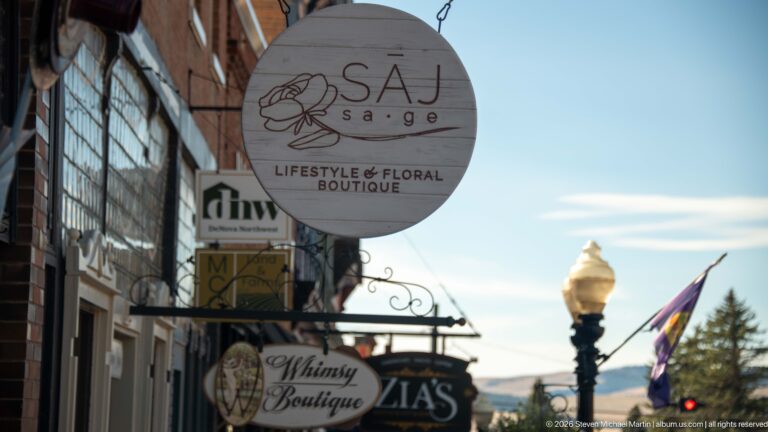 Hanging SAJ Sage boutique sign and other business signs above a Philipsburg sidewalk, with a streetlamp and mountains beyond.