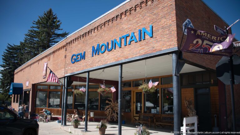 Gem Mountain storefront in a long brick building with blue lettering, covered sidewalk, flags, display windows, and outdoor merchandise.