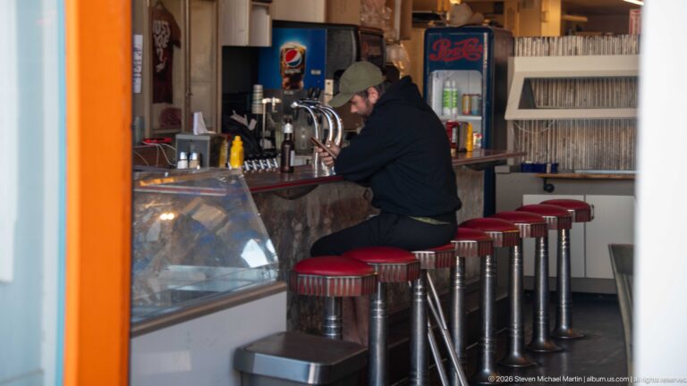 Row of red-topped chrome stools at a counter with beverage taps, coolers, and a worker behind the service area.