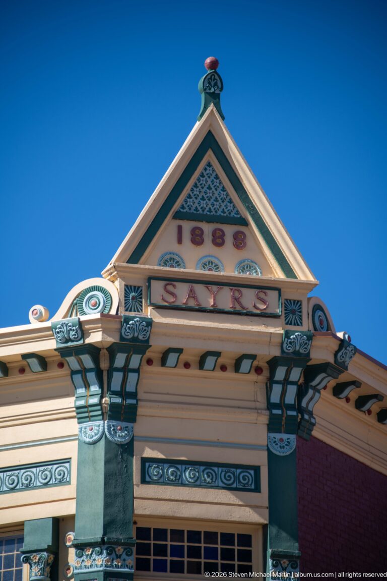 Upward view of an ornate cream-and-green historic building cornice with triangular pediment, brackets, scrollwork, and patterned panels.