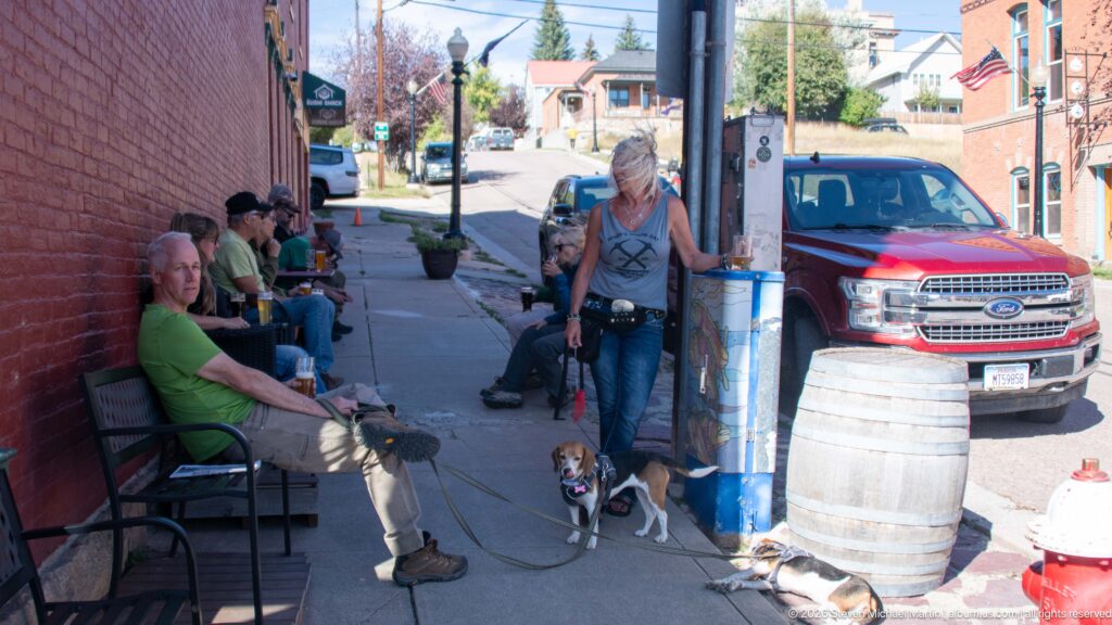 People with beagles gathered along a brick storefront sidewalk beside a barrel, lamppost, parked truck, and uphill street.