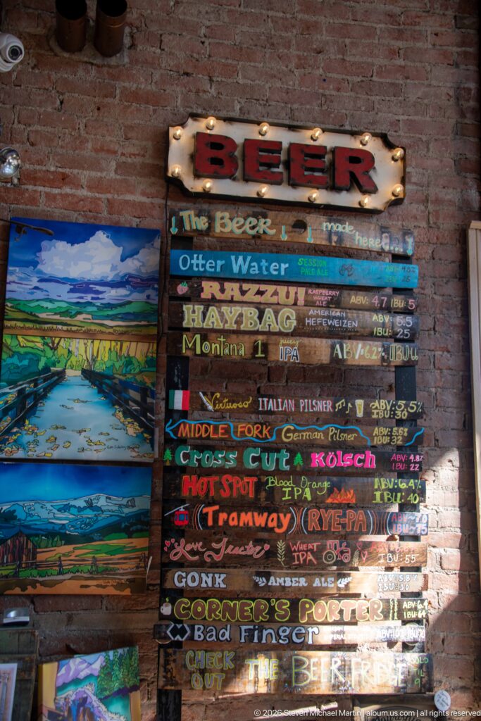 Colorful hand-painted beer menu on stacked wooden slats beside landscape paintings on a brick interior wall.
