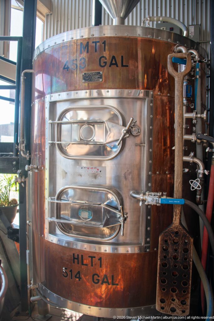 Copper-clad 514-gallon brewery tank with stainless-steel access doors, latches, valves, and a long perforated tool.