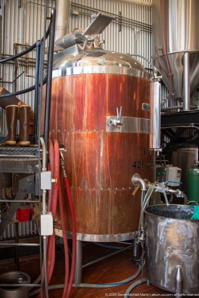 Large copper-clad brewing vessel surrounded by stainless-steel tanks, hoses, valves, and other brewery equipment.
