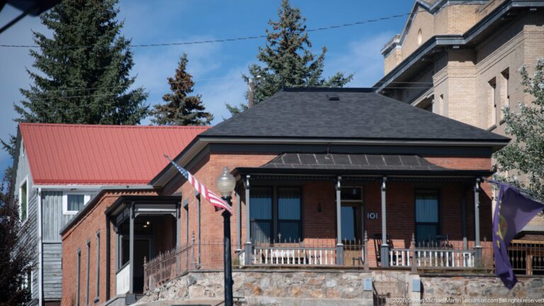 Small historic brick building at number 101 with a hipped roof, front porch, wooden railing, and stone retaining wall.