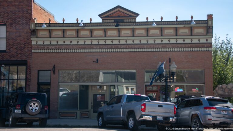 Front view of the J.C. McLeod Building with central pediment, elaborate painted cornice, brick storefront, and vehicles parked in front.