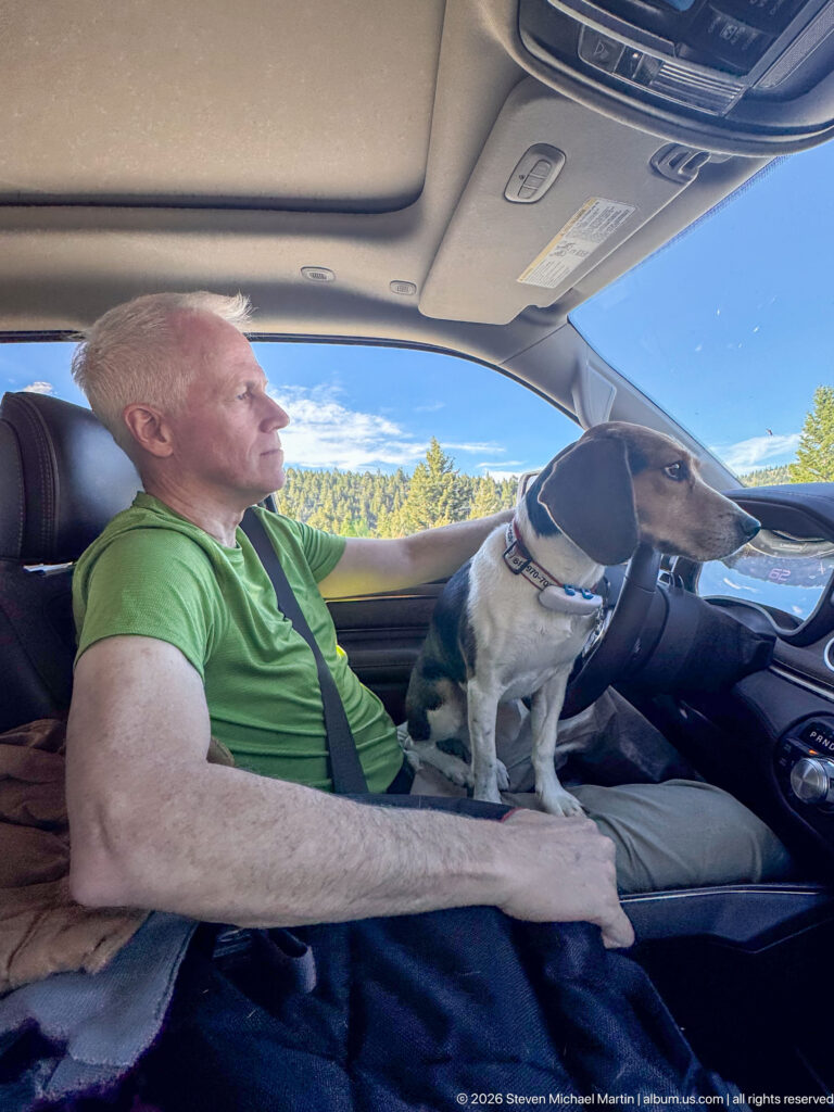 Beagle standing alert on the front seat beside a man in a green shirt, with wooded hills visible outside.