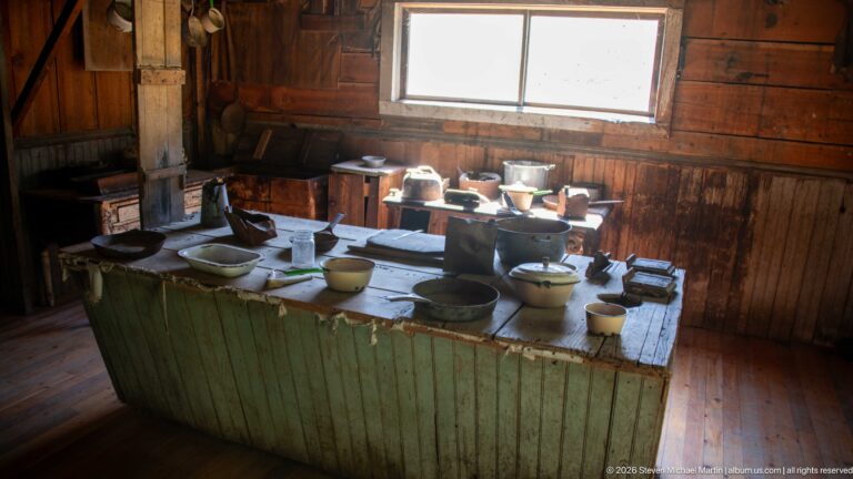 Large green wooden worktable covered with bowls, cups, pots, pans, and enamelware beneath a bright window.