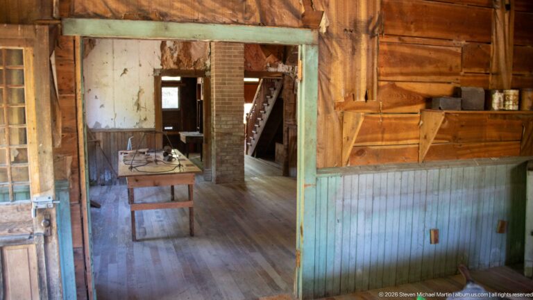 Blue-green paneled service opening framing a table, brick chimney, doorway, and staircase in adjoining wooden rooms.