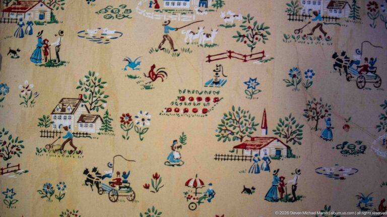 Pale wallpaper patterned with colorful miniature houses, trees, flowers, people, animals, fences, and horse-drawn vehicles.