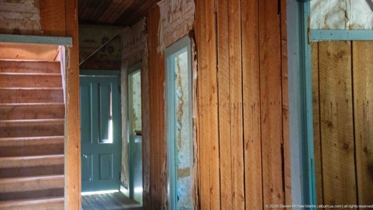 Wooden staircase beside a narrow plank-walled corridor with blue-green doors and bright daylight entering through an open doorway.