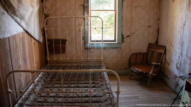 Bare iron bed facing a tall window, with two wooden rocking chairs and extensively peeling wall covering.