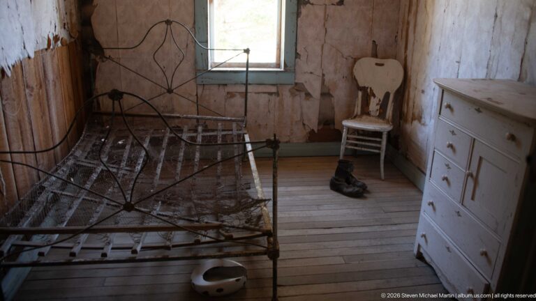 Iron bed beside a white dresser and chair, with boots and a chamber vessel on the floor beneath a small window.