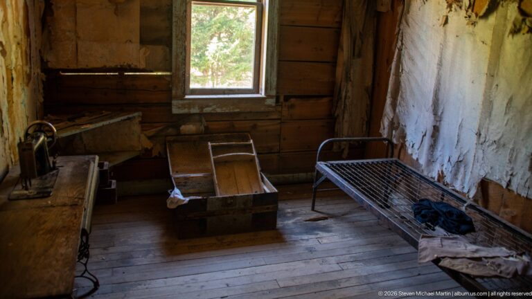 Open wooden trunk beside an iron bed in a small plank-walled room with a window and deteriorated pale wall finish.