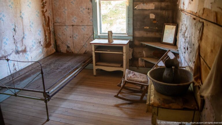 Historic room with iron bed frame, small table, rocking chair, large basin, window, wood floor, and peeling walls.