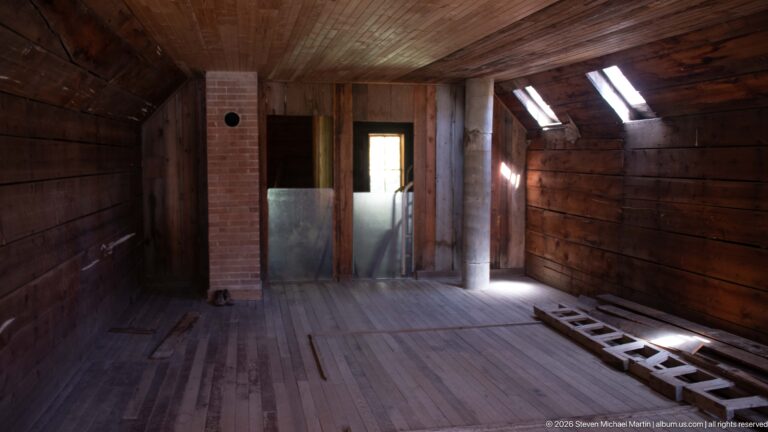 Long wood-paneled room with plank floor, brick chimney, bright roof windows, doorway, and an iron bed frame.