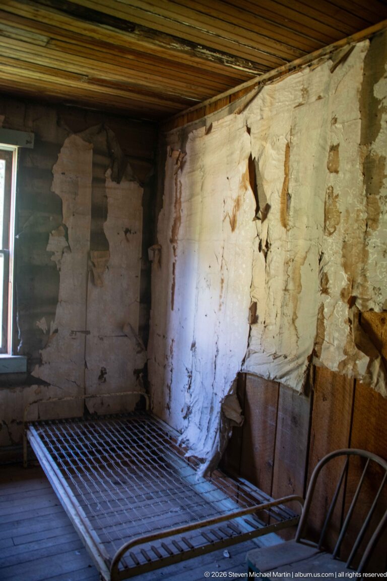 Iron bed frame in a bare wooden-floored room beside a cracked, partially missing plaster wall and small window.