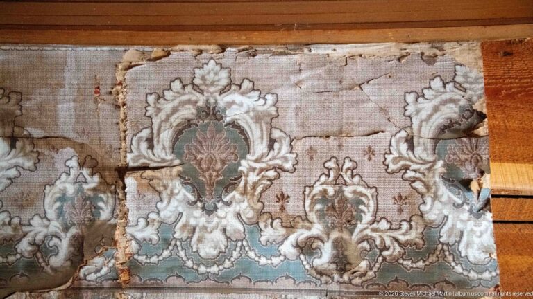 Torn historic wallpaper with large cream, green, brown, and rose ornamental motifs attached to horizontal wooden boards.