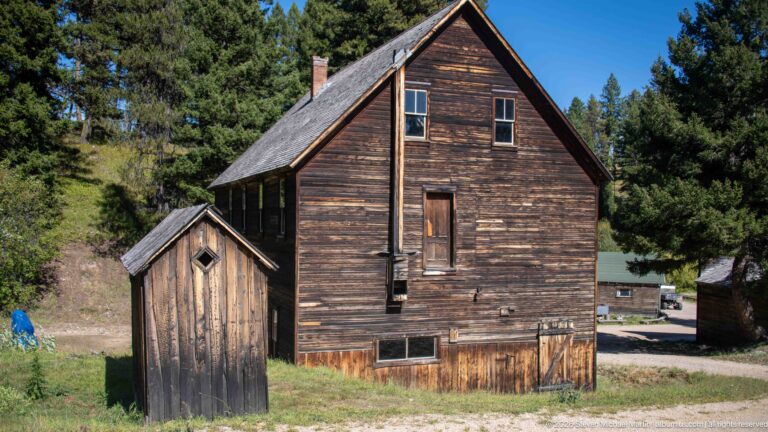 Two-story weathered wooden building with steep gable roof beside a small detached wooden privy in a grassy clearing.