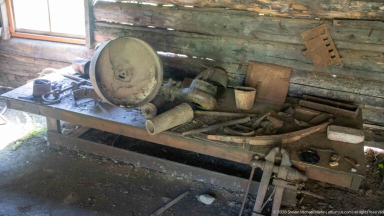 Wooden workbench covered with hand tools, metal containers, hardware, and other artifacts inside a rough log-walled room.