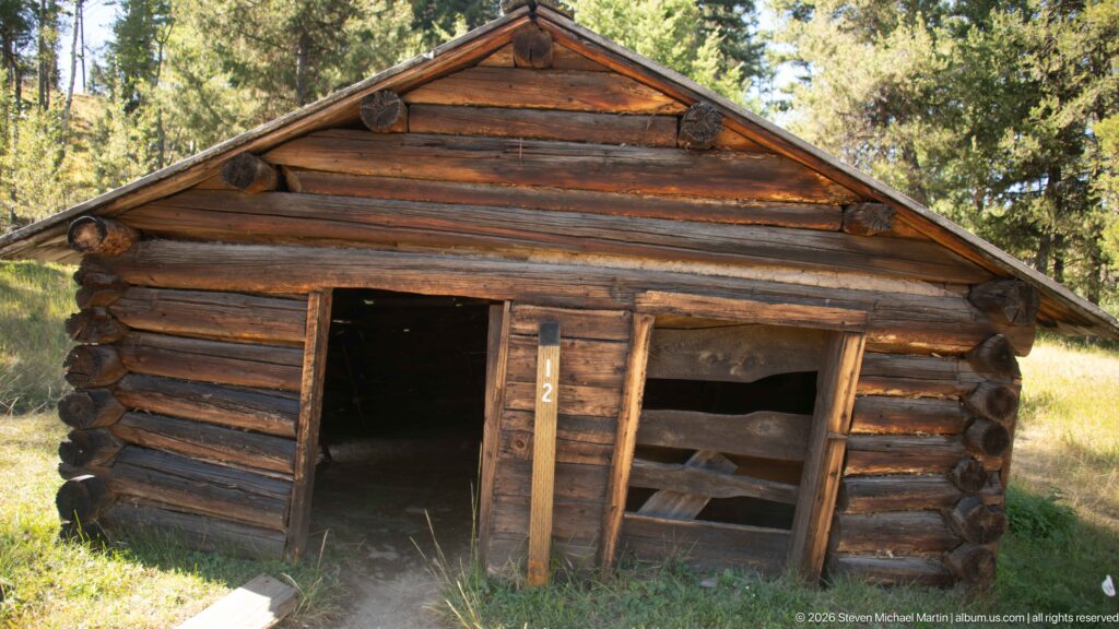 Small weathered log building with an open doorway, wall opening, and projecting log ends in a forest clearing.