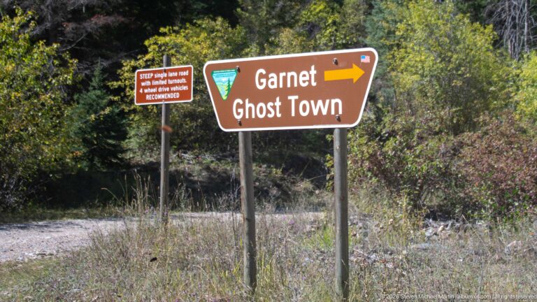 Brown Garnet Ghost Town directional sign with a right-pointing arrow beside a smaller winter warning sign in roadside vegetation.