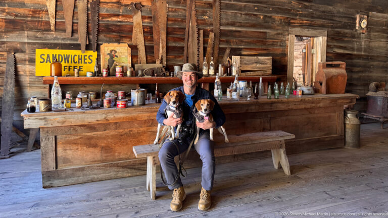 Visitor seated on a wooden bench holding two beagles before a historic bar filled with bottles, tins, and tools.