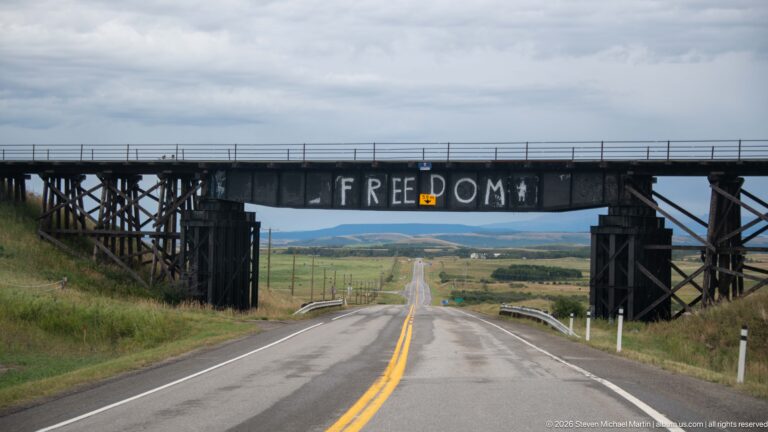 Two-lane highway passing beneath a dark timber trestle painted with the word FREEDOM, with open ranchland and distant hills beyond.