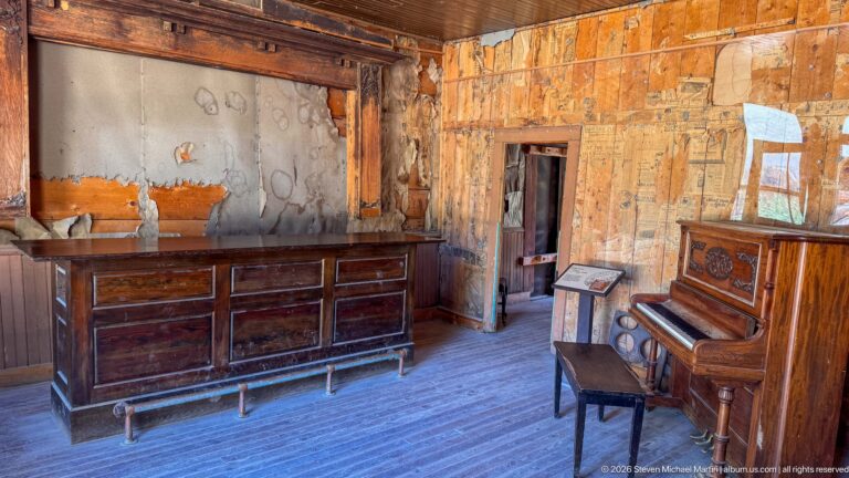 Long wooden bar and upright piano inside a worn historic room with exposed newspaper wall covering and plank floor.