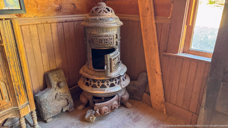 Tall ornate cast-iron stove with pierced decoration and domed top standing beside a window on a wooden floor.