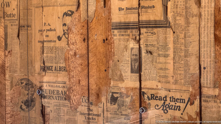Aged newspaper pages pasted over rough wooden boards, with advertisements and headlines visible between tears and nail holes.