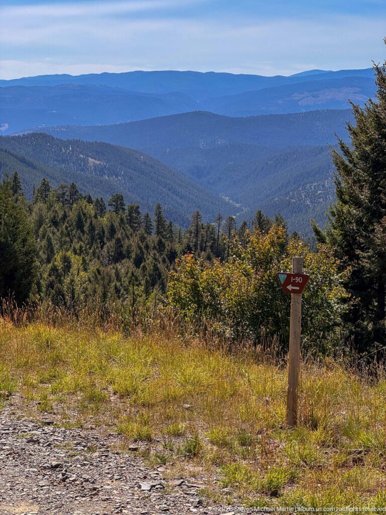 Layered blue and forested mountain ridges viewed from a grassy roadside clearing with a small route marker.