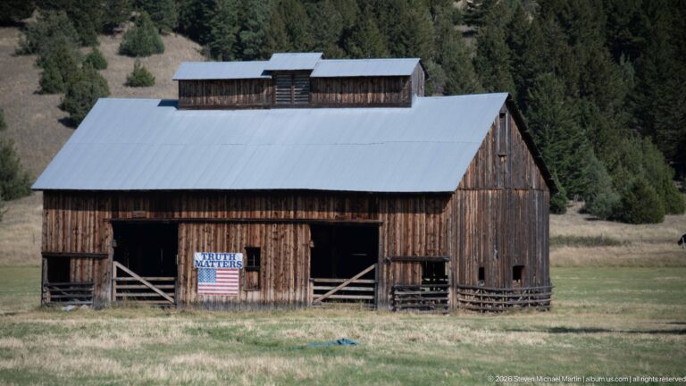 Large weathered wooden barn with a gray metal roof, open bays, fencing, and an American flag in a grassy meadow.