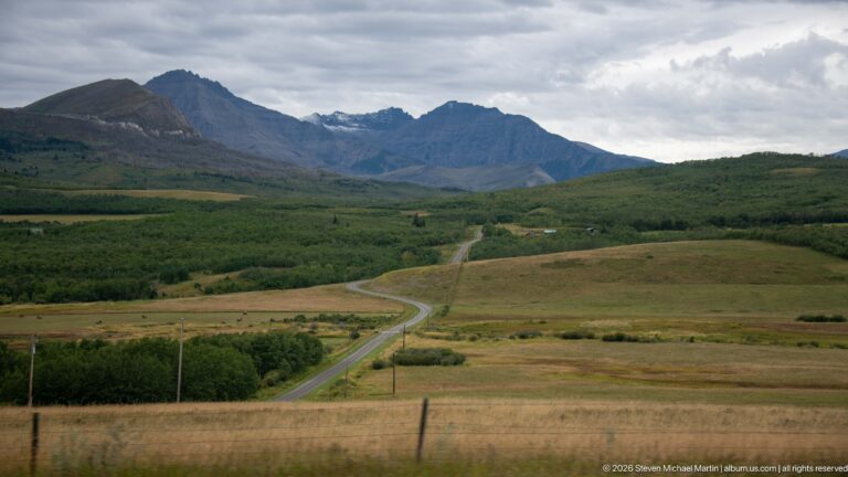 Winding paved road crossing open grassland and wooded foothills toward a rugged mountain range beneath heavy gray clouds.