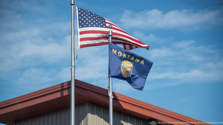 United States and Montana flags flying on adjacent poles above a red-roofed building under a blue sky.