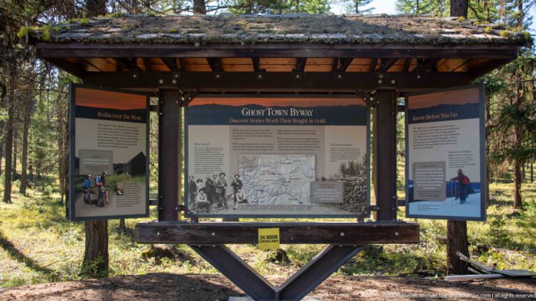 Roofed timber kiosk in conifer forest holding three Ghost Town Byway interpretive panels with maps, history, and visitor guidance.