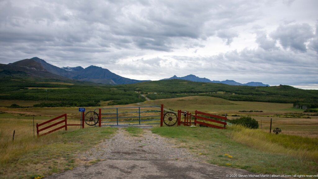 Red-and-blue ranch gate with decorative wagon wheels at the end of a gravel drive, backed by pasture, foothills, and rugged mountains.