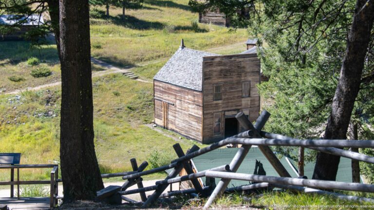 Two-story wooden false-front building seen downhill through tree trunks and a rustic crossed-pole fence.