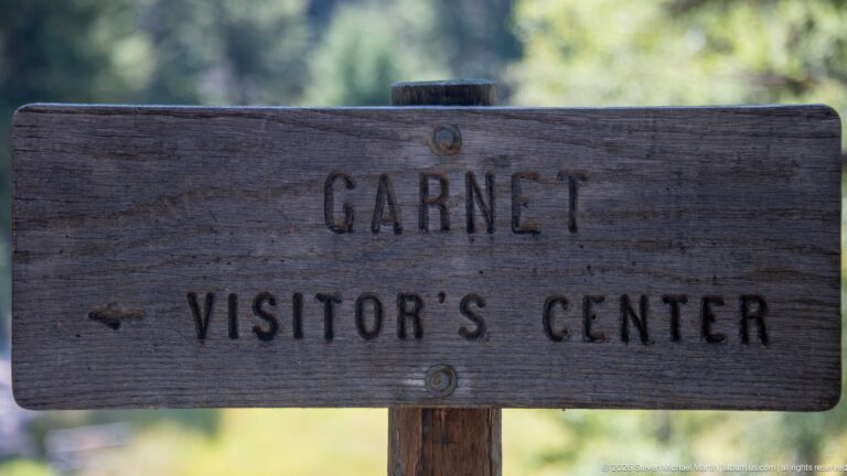 Weathered wooden sign reading Garnet Visitor’s Center with a left-pointing arrow against a blurred forest background.